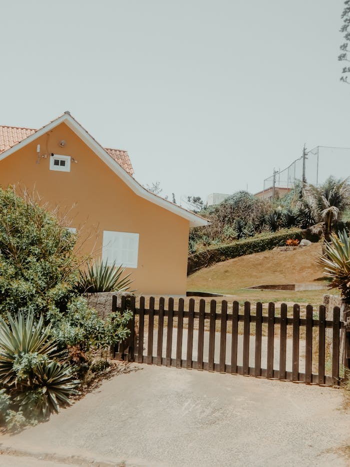 Charming yellow house in a sunny suburban neighborhood with a wooden fence and lush greenery.